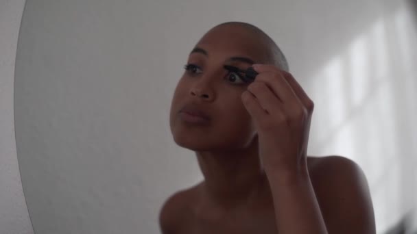 African American female with bald head doing makeup and putting black mascara on eyelashes