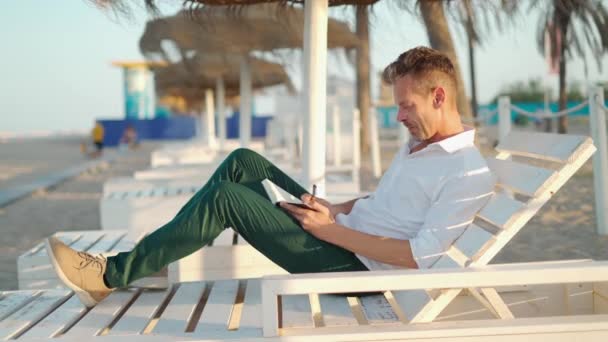 スマートなカジュアルな服の起業家 ラウンジャーに座ってノートブックで執筆しながら海の近くの砂浜の遠隔プロジェクトに取り組んで — ストック動画