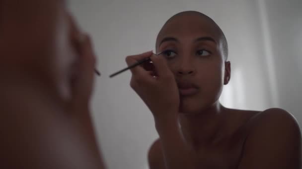 アフリカ系アメリカ人女性の頭がはげ頭で化粧をし 鏡を見ながらブラシで液体アイライナーを適用 — ストック動画
