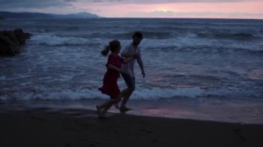Sahilde gün batımında deniz dalgalarıyla eğlenen ve birlikte koşan neşeli, yalınayak romantik çiftin yan görüntülerini izliyoruz.