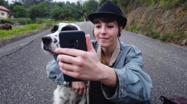 Günlük kıyafetleri ve şapkası olan neşeli genç bir bayan cep telefonuyla yerde oturuyor ve sokakta yürürken köpekle selfie çekiyor.
