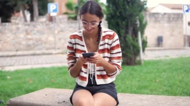 Kentte bankta oturan olumlu genç bir kadın ve ekrana bakarken cep telefonuyla sosyal medyada mesajlaşıyor