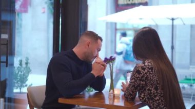Romantik çift, randevu sırasında modern kafeteryadaki masada cep telefonuyla sosyal medyaya göz atarken video izliyor.