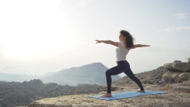 Dans Tanrısı pozisyonunda kayalık dağın tepesinde yoga yaparken bulutlu gün batımına karşı duran esnek kadının alçak açılı yan görüntüsü