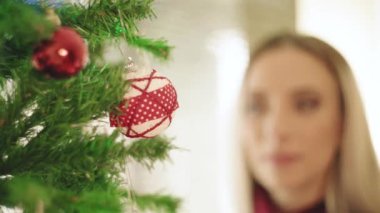 Cheerful young blond female in red sweater hanging decorative ball on Christmas tree while preparing for home celebration