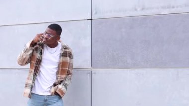 Kareli tişörtlü mutlu siyah adam, sokaklarda yürürken gülümsüyor ve telefona cevap veriyor.