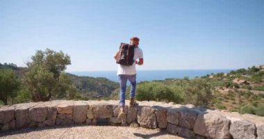 Sırt çantalı yetişkin bir yürüyüşçü taşların üzerinde duruyor ve yaz günü eski bir kasabayla dağ ve deniz manzarasının keyfini çıkarıyor.