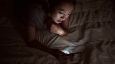 Asyalı bir kadın evde akıllı telefon kullanarak yatakta yatıyor..