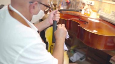Side view of mature male luthier in apron, glasses and protective face mask sitting on chair and holding restored violin while working in workshop