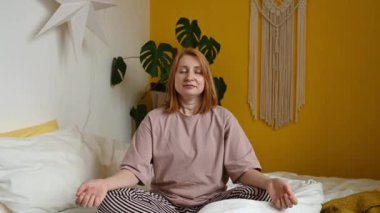 Padmasana yoga pozisyonunda oturan genç kızıl saçlı kadın. Gözleri kapalı ve rahat bir yatakta meditasyon yaparken Mudra elleriyle poz veriyor.