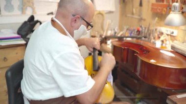 Side view of mature male luthier in apron, glasses and protective face mask sitting on chair and holding restored violin while working in workshop