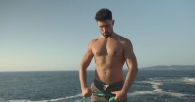 Çıplak vücutlu kaslı erkek vücut geliştirici. Deniz kıyısında duruyor ve yaz aylarında egzersiz yaparken direnç bandıyla egzersiz yapıyor.