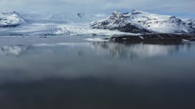 Donmuş göl kenarındaki karla kaplı kayalık dağların nefes kesici manzarası Vatnajokull Ulusal Parkı 'ndaki bulutlu mavi gökyüzüne karşı.