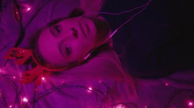 From above of relaxed young female with Christmas reindeer horns and earphones listening to music while lying on bed with glowing garland in dark bedroom with neon illumination