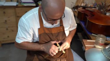 Male master craftsman using professional tool working on violin bridge while building musical instrument in workshop
