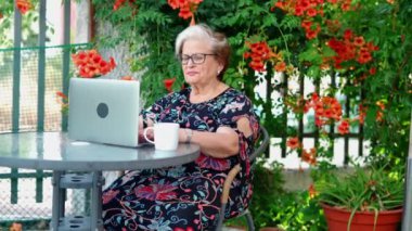 Günlük kıyafetli ve gözlüklü yaşlı bir kadın masada otururken netbook 'un yanında çay tabağı ve bahçede çiçekli yeşil bitkiler üzerinde sörf yapıyor.