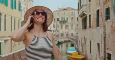 Gülümseyen ve hasır şapkasını çıkartan güneş gözlüklü mutlu kadın Venedik, İtalya 'da kanal üzerinde köprüde akıllı telefonla konuşuyor.