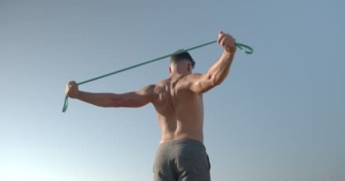 Deniz kıyısında çıplak vücutlu kaslı erkek vücut geliştiricinin arka planında yaz antrenmanları sırasında direnç bandıyla egzersiz yaparken görülüyor.