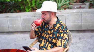 Neşeli İspanyol erkek, boyalı tırnaklı cep telefonuyla mesaj atıyor açık kafedeki masada kokteyl içerken.