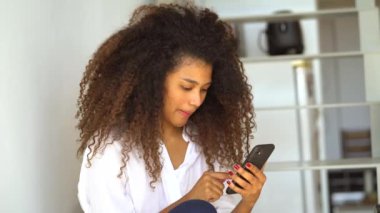 Genç Afro-Amerikalı kadın rahat kıyafetlerle internette cep telefonuyla dolaşırken evde dinleniyor.