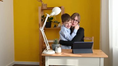 Mavi tişörtlü çocuk evden uzakta çalışırken elinde tabletle masada oturan gözlüklü mutlu anneyi kucaklıyor.