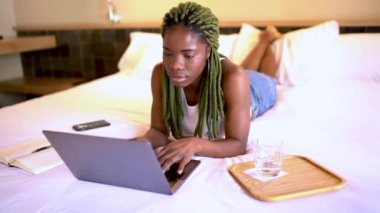Evdeki modern yatak odasında uzaktan çalışırken ve su içerken yatakta uzanan ve netbook 'u karıştıran genç etnik bir kadın.