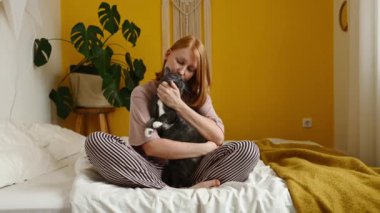 Pijamalı pozitif kızıl kadın bacak bacak üstüne atmış, rahat bir yatakta oturuyor ve beyaz pençeli sevimli kediyi okşuyor.
