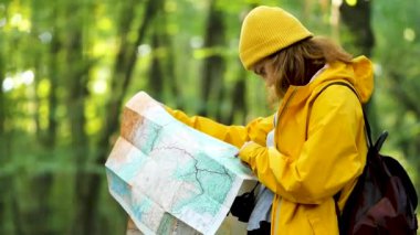 Side view of female explorer in yellow raincoat and with backpack standing in woods and searching for route on paper map
