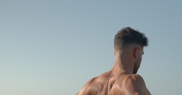 背景色海岸に立って裸の胴を持つ筋肉の男性のボディビルダーと夏のワークアウト中に抵抗バンドと演習を行う — ストック動画