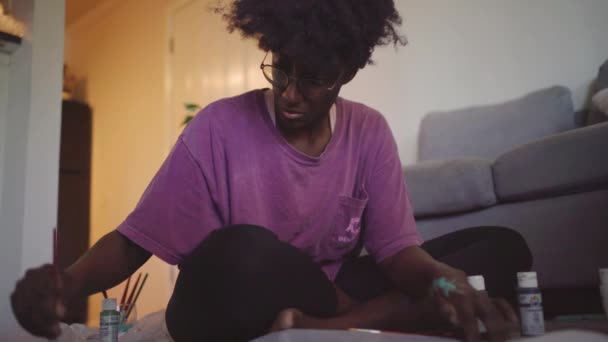 アフロの髪を持つ若い美しいアフリカ系アメリカ人女性 居心地の良いアパートで床に座っている間 紙の上に絵筆でカラフルな絵を描くカジュアルな衣装のアマチュアアーティスト — ストック動画