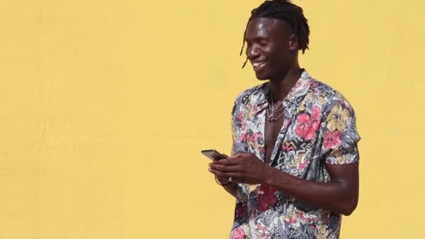 年轻快乐的嬉皮士非洲裔美国男人 留着可怕的头发 穿着时髦的夏季衬衫 有花纹 牛仔裤 背景是黄色的手机 — 图库视频影像