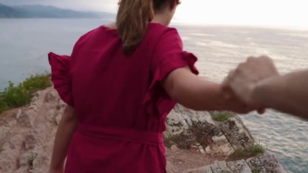 ロマンチックなカップルのバックビューは 日没時に海から荒い岩の崖の上を歩く手を保持しています — ストック動画