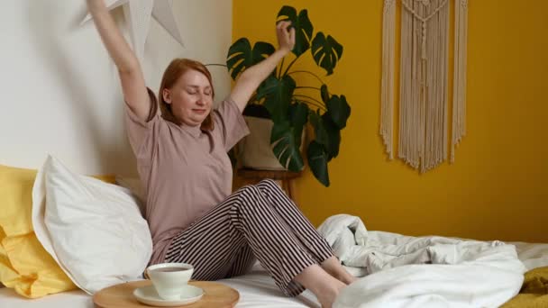 赤毛の裸足の女性は 朝に腕を伸ばして熱い飲料を飲んでベッドに座ってストライプパンツで — ストック動画