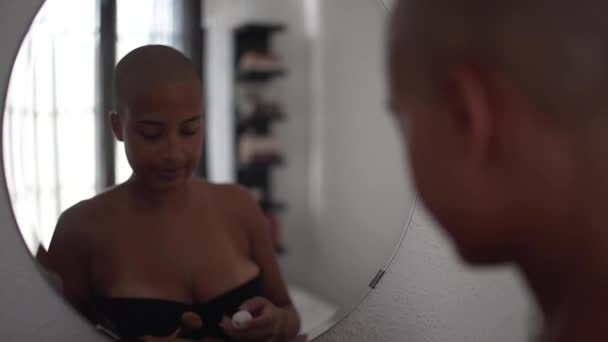 Concentrated African American female with bald haircut looking in mirror and applying foundation with brush while doing makeup