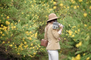 Sıradan giysiler içinde gülümseyen zayıf kadın ve şapka eski video kamera ve deri çantayla kırsalda renkli çiçeklerle çalıların yanında geziniyor ve başka tarafa bakıyordu.