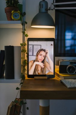 Coronavirus karantinasında bir fotoğrafçıyla görüntülü görüşme yapan sarışın bir gencin portresi. Tablet bir masanın üstünde, bir bilgisayar, birkaç kitap ve bir kamera ile birlikte..