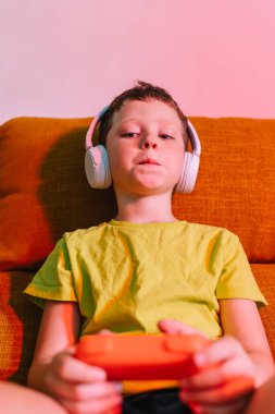 Oturma odasındaki kanepede video oyunu oynarken kulaklıklı, kablosuz kulaklıklı çocuk gözlerini kaçırıyor.