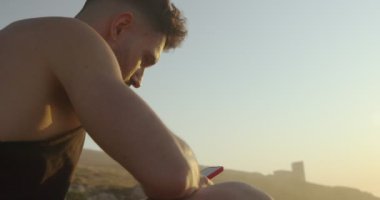 Yazlık giysiler giymiş, deniz kenarındaki kayalarda oturan ve gün batımında cep telefonuyla sosyal medyada mesajlaşan bir erkek.