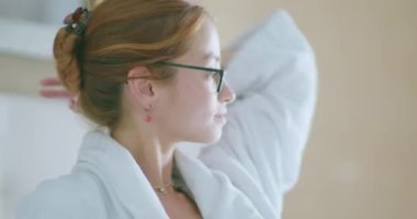 Yumuşak beyaz bornoz ve gözlüklü genç bir kadın açık renk yatak odasında uzun dalgalı kızıl saçlara dokunuyor.