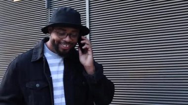 Şapkalı ve gözlüklü neşeli Afrikalı Amerikalı hippi adam sokakta metal duvarın yanında dururken cep telefonuyla konuşuyor.