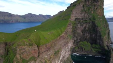 Faroe Adaları 'ndaki bulutlu mavi gökyüzüne karşı yeşil yosunlarla kaplı engebeli uçurum manzarası.