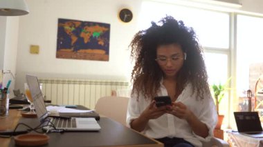 Ciddi bir Afrikalı Amerikalı kadın girişimci ofis masasında oturmuş mesai saatlerinde cep telefonundan mesajlar okuyor.