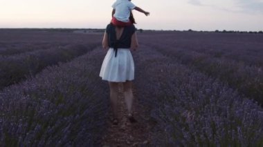 Omuzlarında küçük bir çocuk olan, mor lavanta çiçekleriyle çiçek açan ve günbatımının tadını çıkaran, tanınamayan, sevgi dolu bir annenin arka görünüşü.