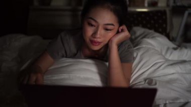 Asyalı bir kadın evde dizüstü bilgisayardan film izlerken yatakta uzanıyor..