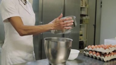 Beyaz üniformalı tanınmayan bir kadın aşçı. Fırın mutfağında hamur işi hazırlarken metal kaseye un karıştırıyor.