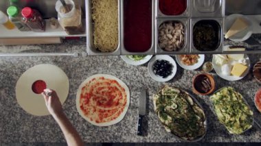 Lokanta mutfağında çeşitli malzemelerle mermer masada pizza pişirirken hamurun üzerine domates sosu dökerek ve yayarak kesilmiş isimsiz şefin üst görüntüsü.