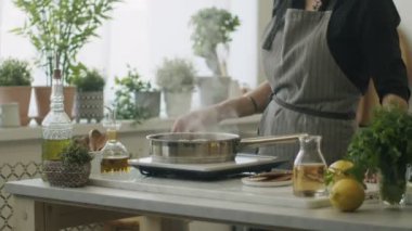 Yemek için mutfak gereçleri ve malzemelerle masada duran, tavada dana inciği pişiren ve tavayı paslanmaz kapakla kaplayan tanınmamış bir kadın.