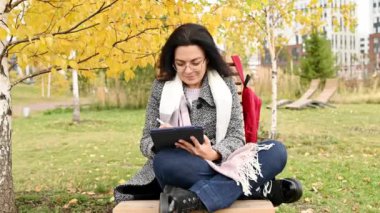 Güz parkında otururken battaniyeye sarılı mutlu genç bir kadın bankta oturuyor ve tableti karıştırıyor.