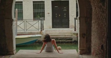 İtalya 'nın Venedik şehrinde haftasonu kanalın yakınındaki bir rıhtımda oturmuş saçları sallayan ve gözlerini kaçıran kadın turistin arka görüntüsü.