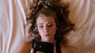 Yukardan gelen mutlu, genç bir bayan yatakta uzanır ve evde dinlenirken akıllı telefondan selfie çeker.